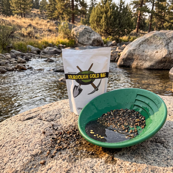 GPAA Sourdough paydirt bag beside a gold pan of gravel on a creekside rock